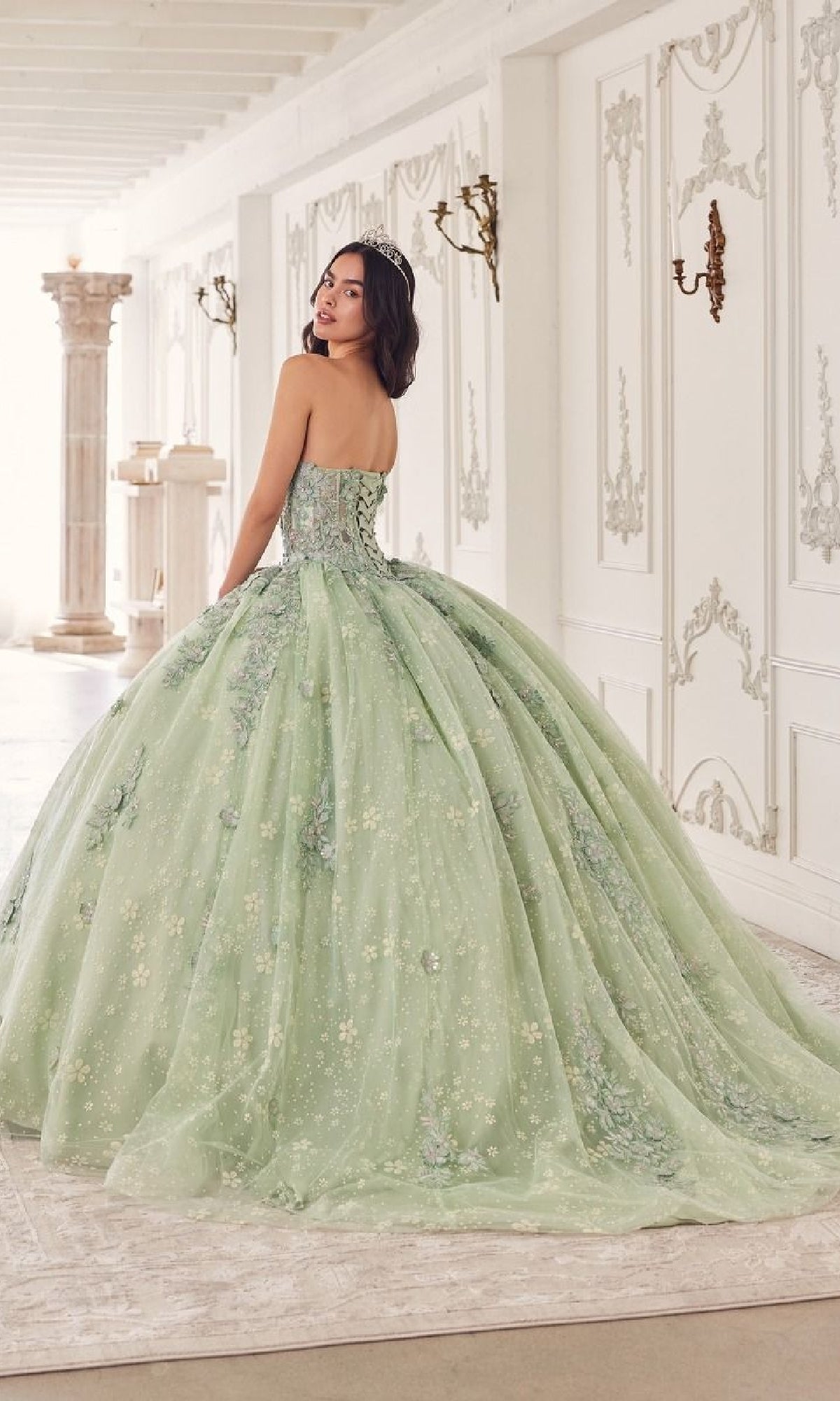 A woman in the Quinceanera Dress 15722 by Ladivine, featuring strapless lace, floral accents, and removable puff sleeves, stands in an elegant white ornate room, looking over her shoulder.